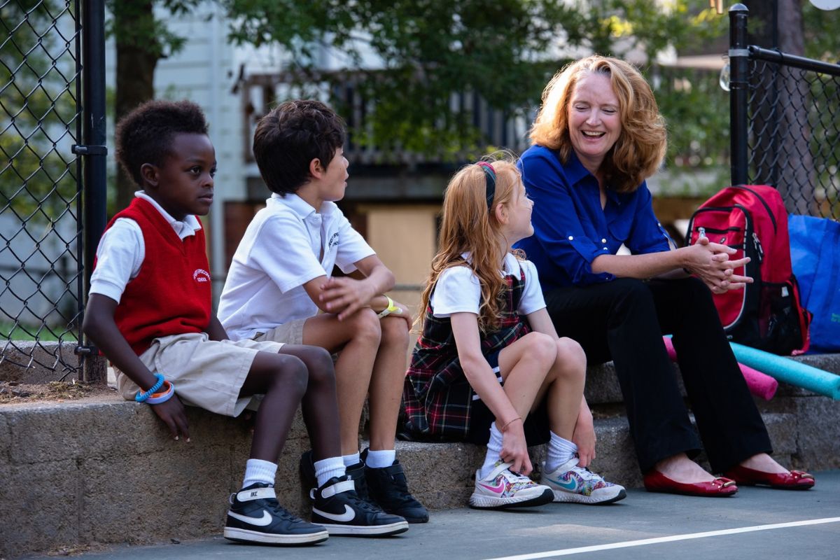 Christ Episcopal School Head of School sits with students in the edge of the playground as they laugh together
